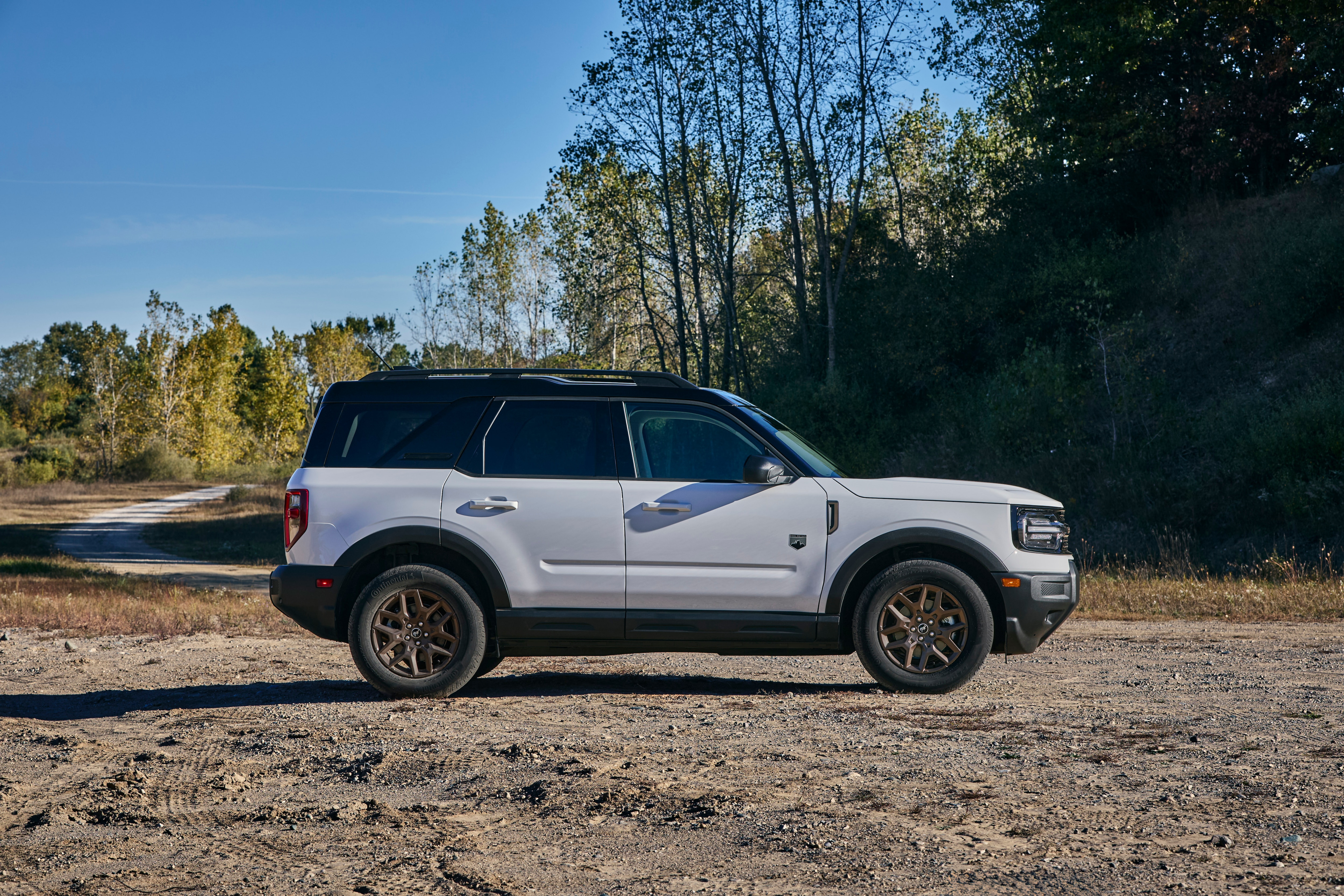 A white Bronco sport with a black top is parked on a gravel lot in a wooded area